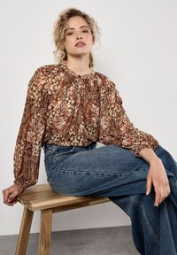 Braun gemusterte Bluse mit gerafftem Ausschnitt und langen Ärmeln, kombiniert mit hochgeschnittenen blauen Jeans, sitzt auf einer Holzbank.