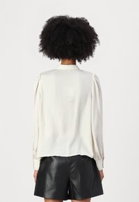 Hellbeige Bluse mit Puffärmeln, strukturiertem Stoff und Kragen, kombiniert mit schwarzen Ledershorts, von hinten betrachtet.