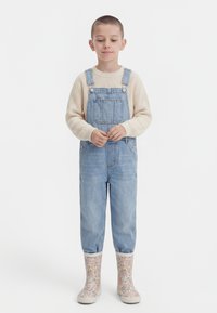 Otrok s kratkimi lasmi, oblečen v bež pulover, denim kombinezo in gumijaste škornje z cvetličnim vzorcem, stoji z rokami prekrižanimi.