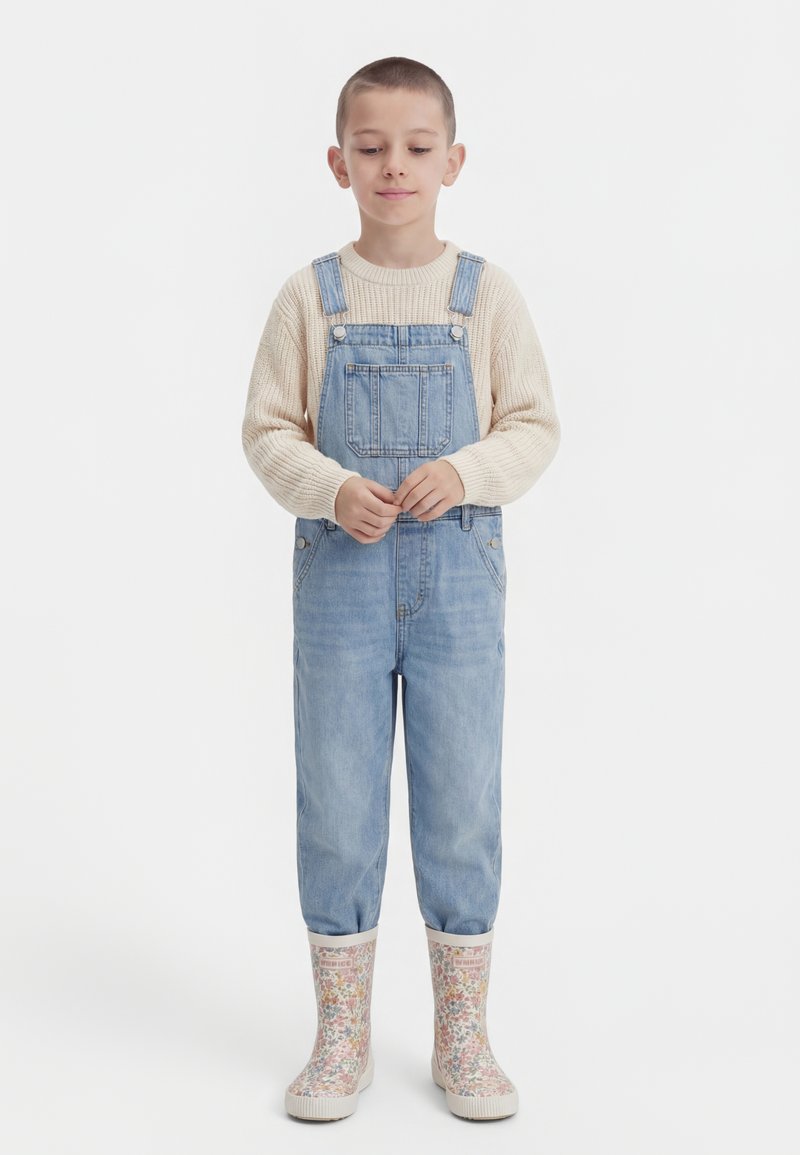 Otrok s kratkimi lasmi, oblečen v bež pulover, denim kombinezo in gumijaste škornje z cvetličnim vzorcem, stoji z rokami prekrižanimi.