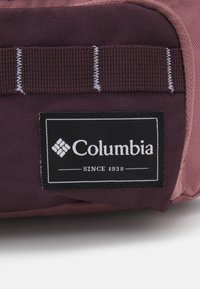 Lähellä kuva viininpunaisesta kangaskassista, jossa on musta suorakulmainen Columbia-brändin etiketti ommeltuna etutaskuun.