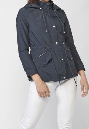 Kvinde iført navyblå hættetrøjejakke med snore, lynlåse og knaplukning på lommerne, kombineret med hvide bukser, stående mod en ensfarvet baggrund.