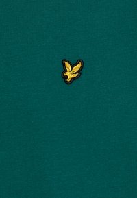 Teal cotton fabric featuring a small embroidered yellow bird logo outlined in black, placed centrally. The texture is smooth with ribbing.