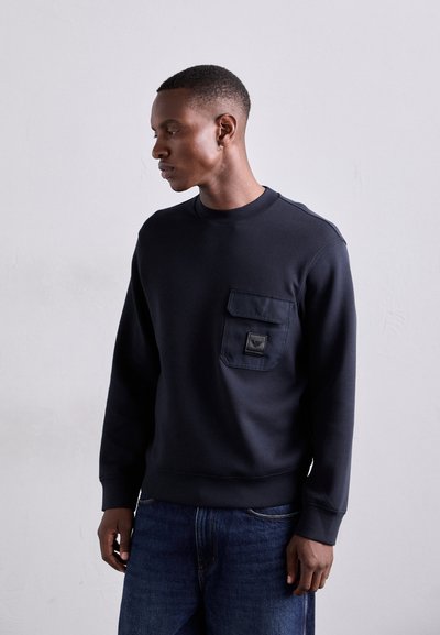 Tamsiai mėlynas palaidinė su apvalia iškirpte, priekyje esančia kišene ir subtiliu logotipo ženkleliu. Minkšta tekstūra ir raištelių rankovės, derinamas su mėlynais džinsais.