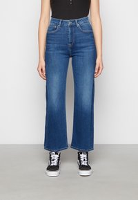 Jeans di denim blu a vita alta con taglio a gamba larga, caratterizzati dal classico stile a cinque tasche e una finitura pulita. Indossati con sneaker nere.