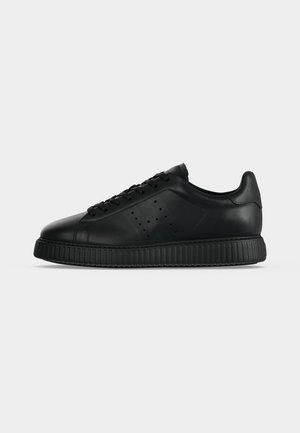 Μαύρο sneaker χαμηλού ύψους με διάτρητο λογότυπο στο πλάι, κορδόνια μπροστά και χοντρή ραβδωτή σόλα, εμφανίζεται από το πλάι σε ανοιχτό φόντο.