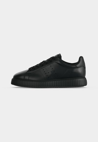Μαύρο sneaker χαμηλού ύψους με διάτρητο λογότυπο στο πλάι, κορδόνια μπροστά και χοντρή ραβδωτή σόλα, εμφανίζεται από το πλάι σε ανοιχτό φόντο.
