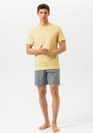 Jonge man staat op blote voeten en draagt een lichtgeel T-shirt en een korte broek met patroon tegen een effen witte achtergrond.