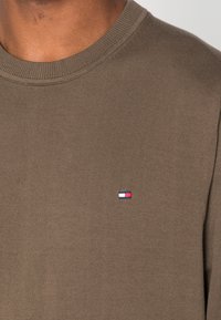 Bruine katoenen trui met ronde hals, geribbelde textuur en aansluitende kraag; heeft een klein geborduurd logo in rood, wit en blauw op de borst.