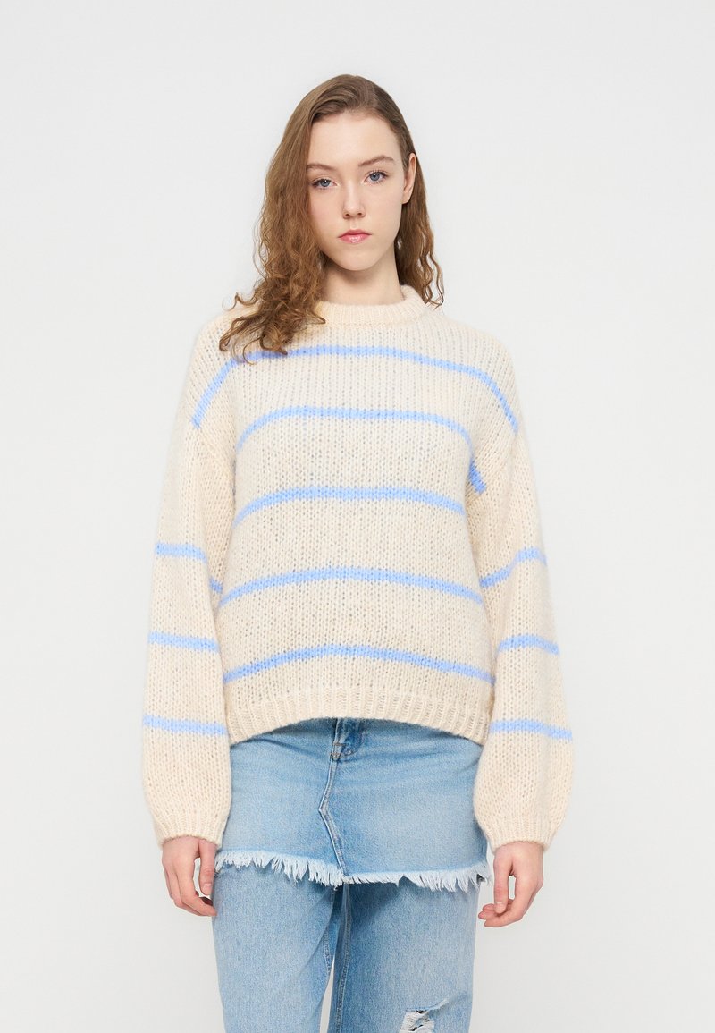 Ανοιχτό μπεζ πλεκτό πουλόβερ με μπλε ρίγες, φαρδιά μανίκια και ριμπ τελείωμα. Συνδυασμένο με σκισμένο ανοιχτό μπλε τζιν.