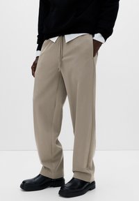 Bézs nadrág puha, texturált anyagból, laza szabással, elasztikus derékrésszel, húzózsinórral és oldalsó zsebekkel, fekete cipővel viselve.