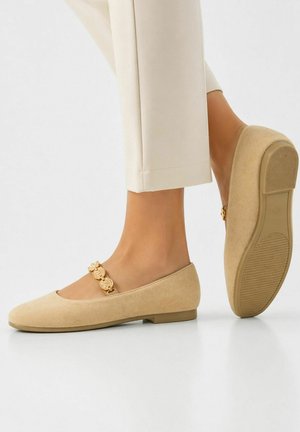 Beige suède platte schoenen met gouden decoratieve riem, gedragen met crèmegekleurde broek, waarbij het profiel van de zool op één schoen zichtbaar is.
