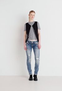 Un gilet sans manches noir sur un t-shirt gris, associé à un jean bleu déchiré et des bottines noires à la cheville. Le modèle se tient contre un fond uni.