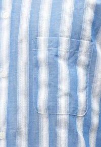 Zils un balts svītraina krekls no viegla auduma, ar krūšu kabatu, nelielām pogām un smalku tekstūru svītru rakstā.