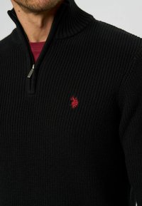 Črni rebrasti pulover z visokim ovratnikom in četrtinskim zadrgo. Vsebuje majhen rdeč vezen logotip na prsih. Tkanina deluje teksturirano.