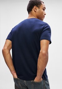 Homem com cabelo curto e encaracolado, vestindo uma t-shirt de manga curta azul-marinho e jeans escuros, com as mãos nos bolsos de trás, de costas para a câmera.