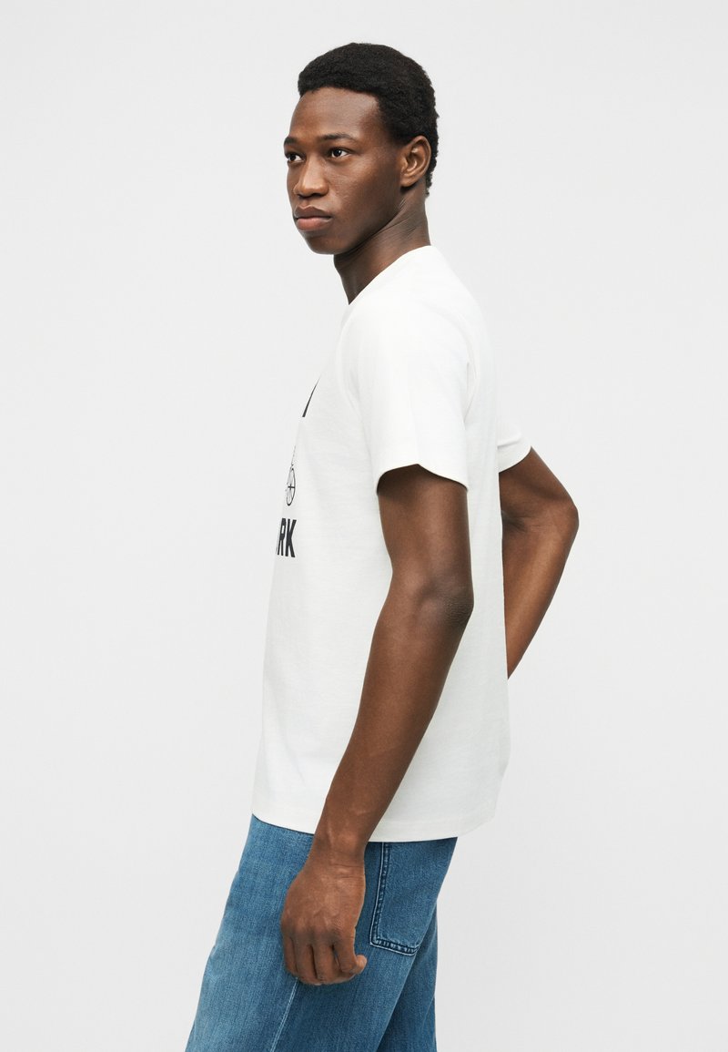 Jeune homme debout de profil portant un t-shirt blanc à motifs et un jean bleu, sur un fond clair uni.