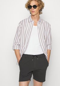 Gestreepte button-up blouse in beige en wit, gedragen over een witte tanktop. Gecombineerd met donkergrijze shorts met elastische tailleband en trekkoord.