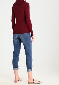 Donna che indossa un maglione con cappuccio color bordeaux, jeans blu risvoltati e scarpe basse beige senza lacci, in piedi davanti a uno sfondo chiaro e semplice.
