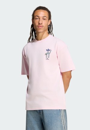 Man met lange dreadlocks die een lichtroze t-shirt draagt met een kleine cartoonkat en het Adidas-logo op de borst, gecombineerd met lichtblauwe jeans.