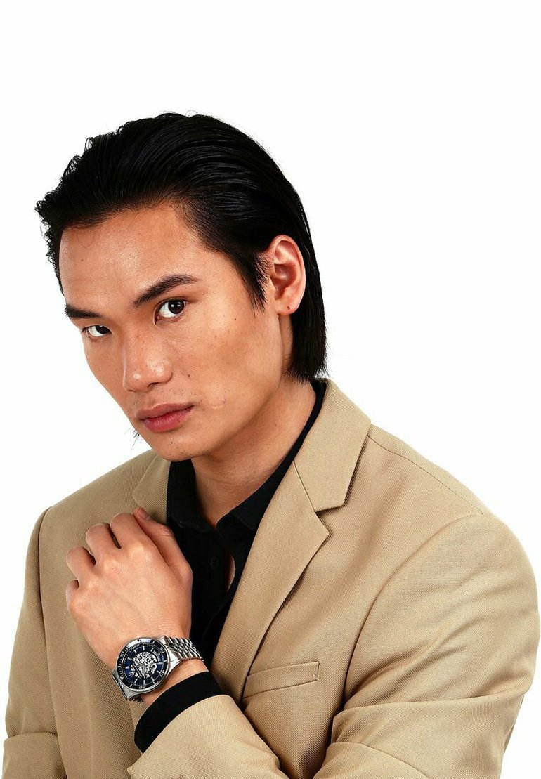 Silver watch with a black dial and metal band, worn on the wrist of a person in a light beige blazer and dark shirt. Simple and modern design.