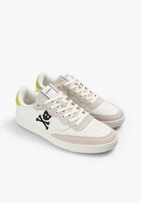 Zapatillas blancas y beige con el logo de calavera y huesos cruzados en negro en los lados, cordones blancos y toques de color verde neón en la zona del talón.