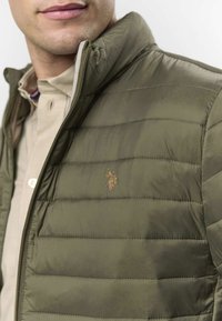 Vestă matlasată olive green cu textură matlasată, închidere cu fermoar și logo brodat auriu. Purtată peste o cămașă bej deschis cu guler.