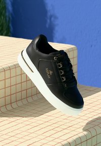 Czarne skórzane sneakersy z białą podeszwą, z zapięciem na sznurówki, złotymi akcentami i logo marki po boku. Gładka tekstura i zaokrąglony kształt.