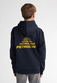 Námořnická mikina s velkým žlutým logem "PETROL IND" na zadní straně, s klokaní kapsou a nastavitelnou kapucí. Měkký materiál z bavlněné směsi.