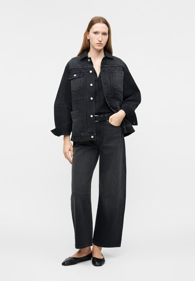 Γυναίκα που φοράει μαύρο τζιν μπουφάν υπερμεγέθους, μαύρο πουκάμισο, μαύρο τζιν παντελόνι με φαρδιές μπλούζες, μαύρη ζώνη και μαύρες μπαλαρίνες, που στέκεται μπροστά σε απλό φωτεινό φόντο.