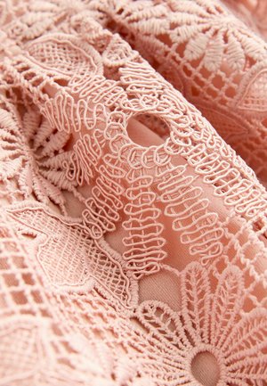 Tissu rose brodé présentant des motifs floraux et géométriques complexes, avec des textures variées et des détails en tricot ouvert.