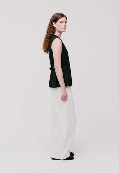 Schwarze ärmellose Weste mit strukturiertem Schnitt und Knöpfen, kombiniert mit weißen weiten Hosen. Das Modell steht vor einem schlichten Hintergrund.