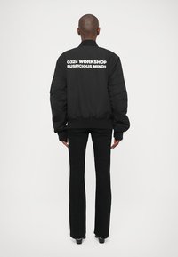 Άτομο που στέκεται με πλάτη στην κάμερα, φορώντας ένα μαύρο μπόμπερ τζάκετ με άσπρο κείμενο "032c WORKSHOP SUSPICIOUS MINDS" και μαύρο παντελόνι.