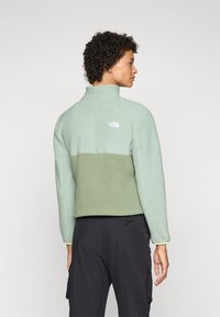 Groene fleecjas met een hoge kraag, met een tweekleurig ontwerp: licht mintgroen bovenaan, olijfgroen daaronder. Klein logo op de achterkant.