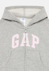 Sivá mikina na zips vyrobená z mäkkého materiálu, so veľkým ružovým logom "GAP" na prednej strane a kapucňou s fleecovou podšívkou. Dve predné vrecká.