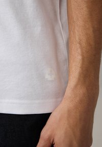 T-shirt bianco in cotone con un sottile logo trasparente sulla parte bassa, abbinato a jeans in denim scuro. Texture liscia e vestibilità casual.