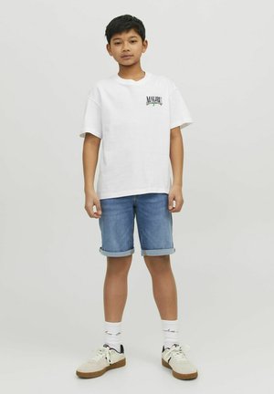 Valkoinen puuvillapaita, jossa on pieni vihreä ja sininen logo, yhdistettynä käärittyihin farkkushorts-t. Jalkineisiin kuuluvat valkoiset lenkkarit, joissa on beige yksityiskohtia.