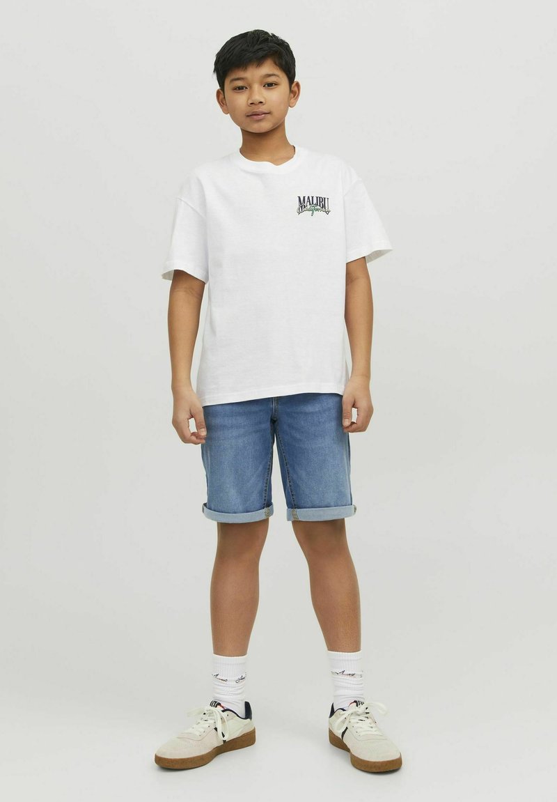 Camiseta de algodón blanca con un pequeño logo verde y azul, combinada con pantalones cortos de mezclilla enrollados. Footwear incluye zapatillas blancas con acentos en beige.