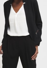 Černý blazer s otevřeným předním dílem, bez knoflíků, se zipovým detailem. Pod ním bílá blůza s V-výstřihem, doplněná černými kalhotami na míru.