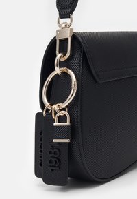 Zwarte leren handtas met textuur en gouden hardware, voorzien van twee zwarte labels, waarvan er één is gegraveerd met "1981," bevestigd aan een gouden ring nabij de sluiting van de schouderriem.