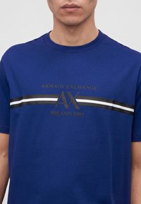 Άνδρας που φοράει μπλε μπλούζα Armani Exchange με μαύρες και λευκές ρίγες και το κείμενο "ARMANI EXCHANGE AX MILANO 1991" στο στήθος.