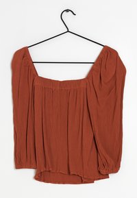 Blouse couleur rouille avec encolure carrée, manches longues bouffantes et tissu texturé, suspendue à un crochet noir sur un fond blanc.