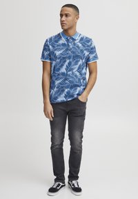 Kortärmad polo skjorta i ljusblått med palmbladsmönster, kombinerad med mörkgråjeans och svarta sneakers med vita detaljer.