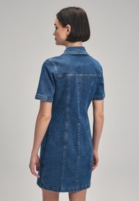 Robe en denim bleu foncé avec un col, des manches courtes et une silhouette ajustée. Présente des coutures visibles et un dos sans couture.