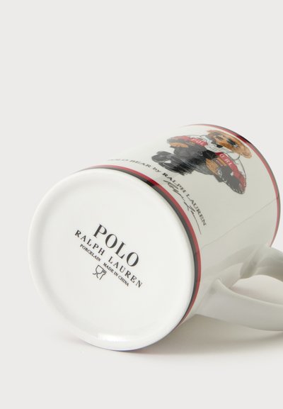 Λευκή πορσελάνινη κούπα Polo Ralph Lauren ξαπλωμένη στο πλάι με σχέδιο αρκούδας και κείμενο της μάρκας στον πάτο.