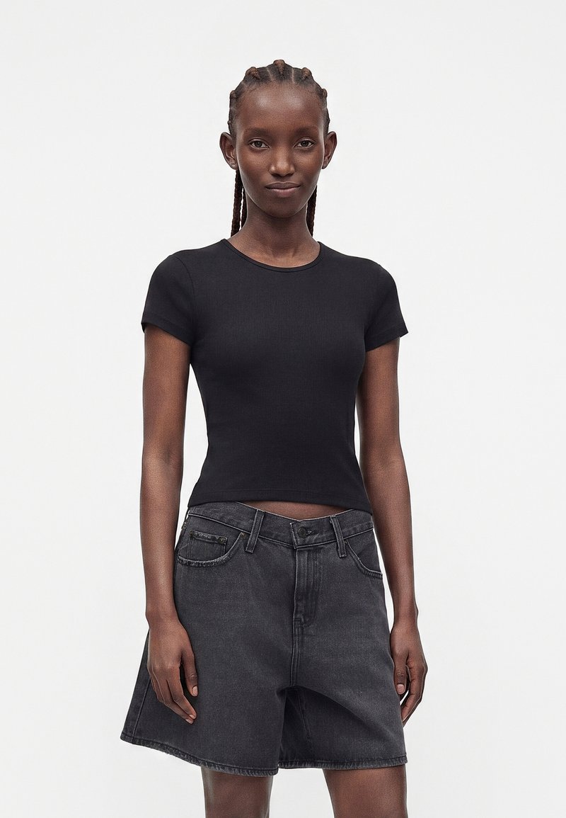 Young woman with braided hair wearing a fitted black short-sleeve top and loose black denim shorts against a plain light background.