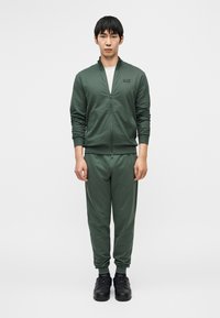 Σκούρο πράσινο φόρμας από μαλακό ύφασμα. Ζακέτα με φερμουάρ και ψηλό γιακά, ελαστικές μανσέτες και αντίστοιχο  στενό παντελόνι. Μαύρα αθλητικά παπούτσια.