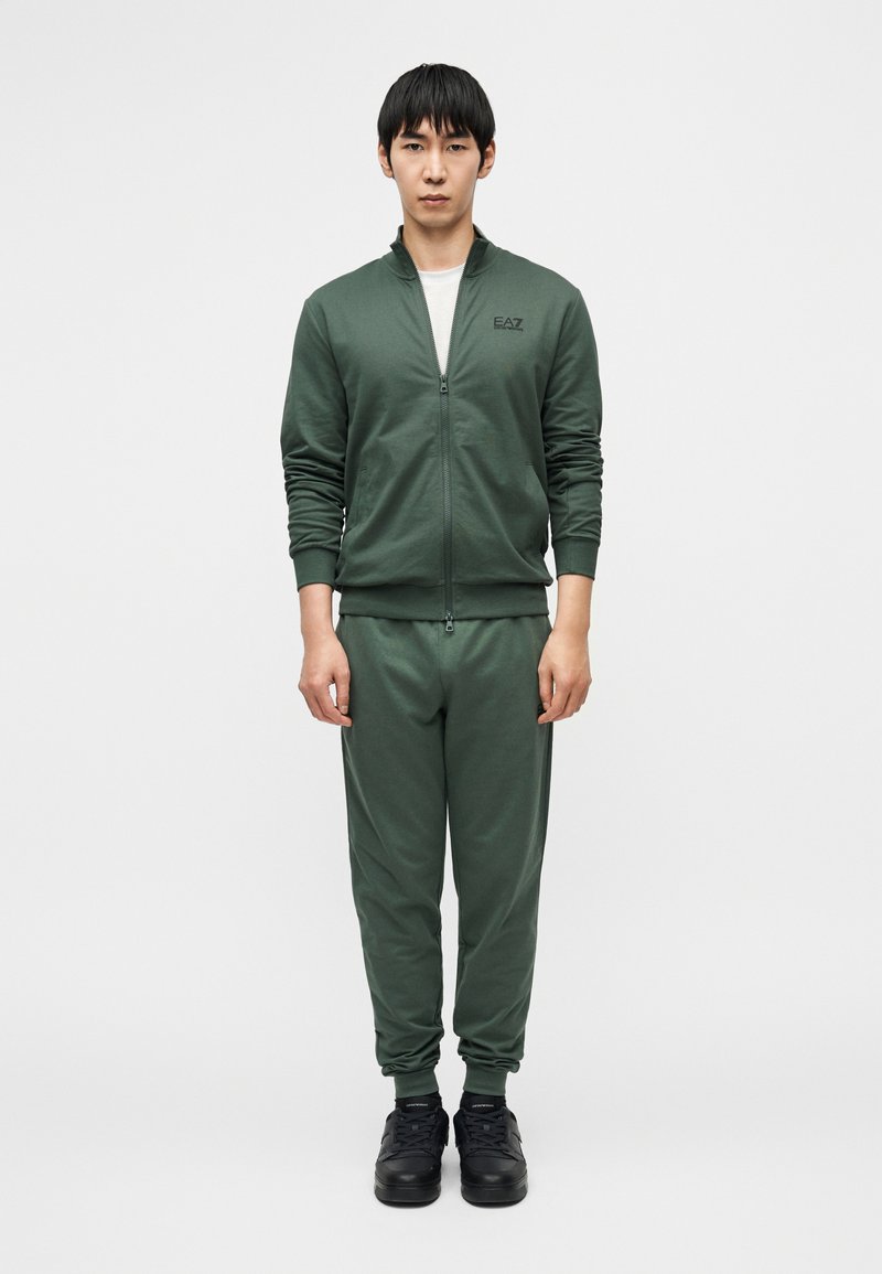 Σκούρο πράσινο φόρμας από μαλακό ύφασμα. Ζακέτα με φερμουάρ και ψηλό γιακά, ελαστικές μανσέτες και αντίστοιχο στενό παντελόνι. Μαύρα αθλητικά παπούτσια.