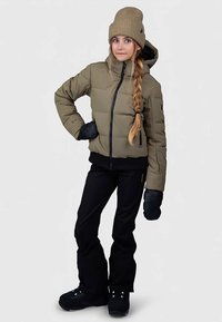 Casaco acolchoado verde-oliva com um fecho preto, combinado com calças e luvas pretas, apresentando um gorro de lã em tom bege e tranças.