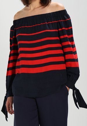 Kobieta ubrana w czarny i czerwony, poziomo paskowany top odsłaniający ramiona z długimi rękawami związanymi na nadgarstkach, w połączeniu z ciemnymi spodniami.